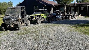A gravel driveway with four off-road vehicles parked in front of a wooden building. - Controlled Chaos Basecamp - centered between Jackson & Clark County Trail System (Merrillan)