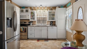 Une cuisine avec des meubles blancs, un réfrigérateur en acier inoxydable, un four à micro-ondes et une cuisinière. Il y a une fenêtre avec des rideaux et une lampe sur le côté droit.