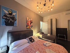 A modern hotel room with a large bed, a headboard, a bedside table with a lamp, a wardrobe, and a mirror.