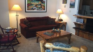 A living room with a red sofa, wooden coffee table, and a rocking chair. There are two lamps, a framed painting, and a bookshelf with a laptop.