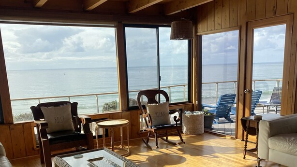 Una habitación amplia con vigas de madera, ventanas grandes que ofrecen vistas al mar, una mesa de centro de vidrio y asientos cómodos.