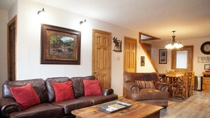 A cozy living room with a brown leather sofa and a wooden coffee table, a wooden dining table with chairs, and a kitchen area with a clock and a sign.