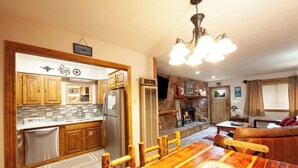 A kitchen with wooden cabinets, a stainless steel refrigerator, and a dining table with chairs. There is a fireplace in the living area with a stone surround.