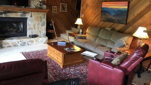 A cozy living room with a stone fireplace, a television, a sofa, a red leather chair, and a painting of a sunset. - FALLING WATERS ~ 2b/2b SPECTACULAR VIEW HANGING OVER FALLS, UPSCALE FURNISHINGS. (Lake Toxaway)