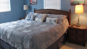 Un dormitorio con cama, mesita de noche, ventilador de techo y dos ventanas con persianas.