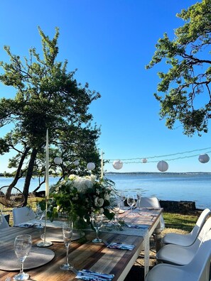 An outdoor dining setup with a view of the water, a wooden table, white chairs, and a string of paper lanterns. - Waterfront Cottage on the Beach In Beautiful Dorset Countryside Sleeps 12 (Dorset)