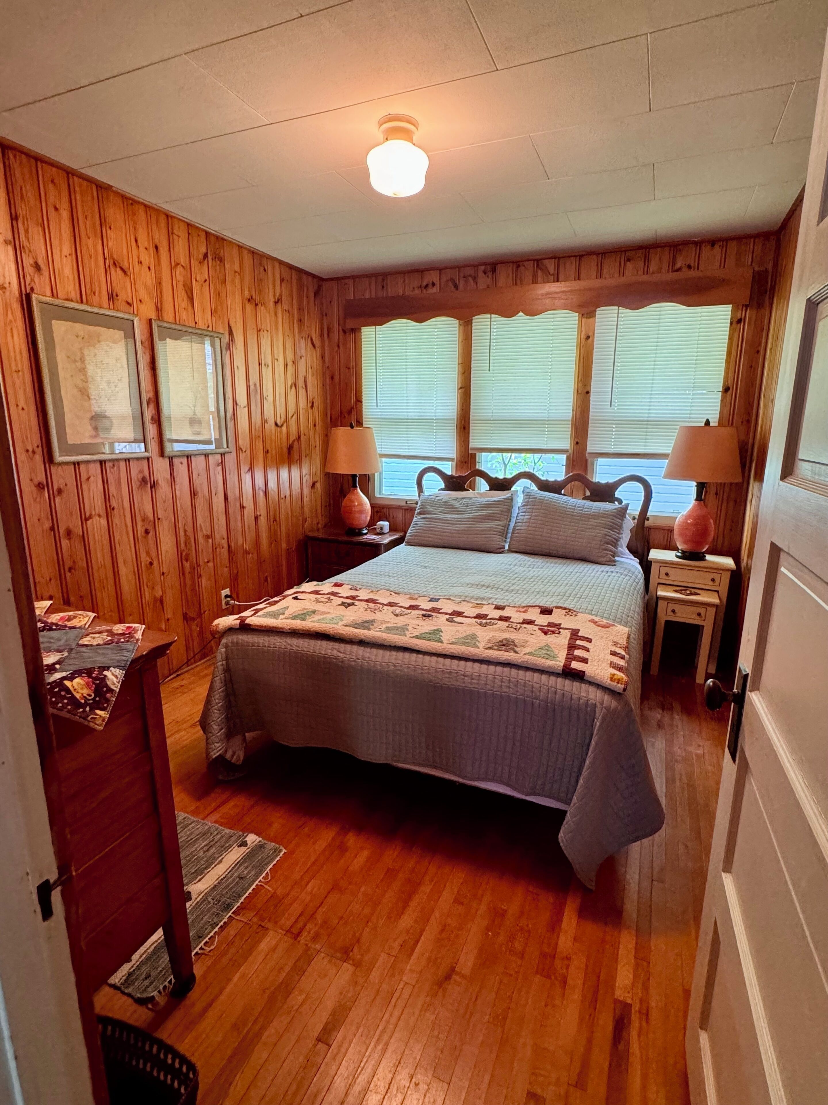 A bedroom with wooden paneling, a bed with a quilt, two bedside lamps, and a window with blinds.