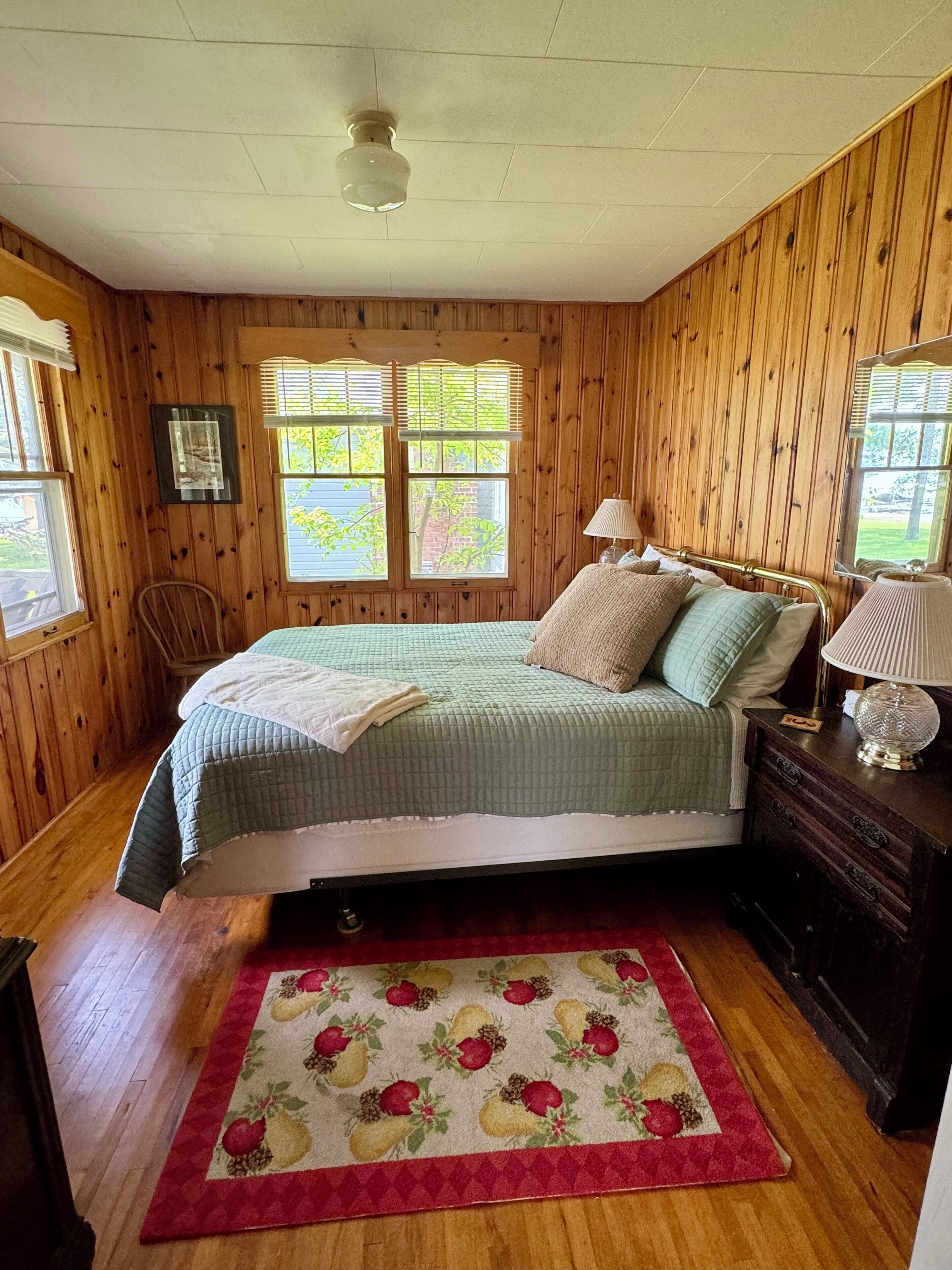 Un dormitorio con paneles de madera, una cama, una mesita de noche, una silla y una ventana con persianas.