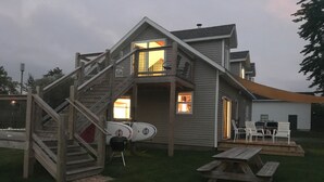 A house with a wooden staircase leading to a balcony, a covered patio area, and a surfboard leaning against the house. - The Narrow’s Yacht Club Emilia’s Loft (Lake Leelanau)