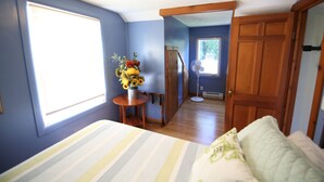 A bedroom with a bed, a wooden door, a window, and a small table with a vase of flowers. - The Narrow’s Yacht Club Emilia’s Loft (Lake Leelanau)