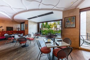 A modern restaurant with a mix of indoor and outdoor seating, featuring a unique ceiling design and a view of tropical plants.