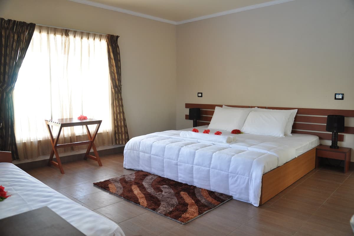 Quarto de hotel com uma cama grande, uma mesinha de cabeceira, uma pequena mesa e uma janela com cortinas.
