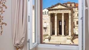 A room with a marble table, a bottle of champagne, and two glasses. A window reveals a building with columns and a clear sky. - Augustus Forum View (Pula)