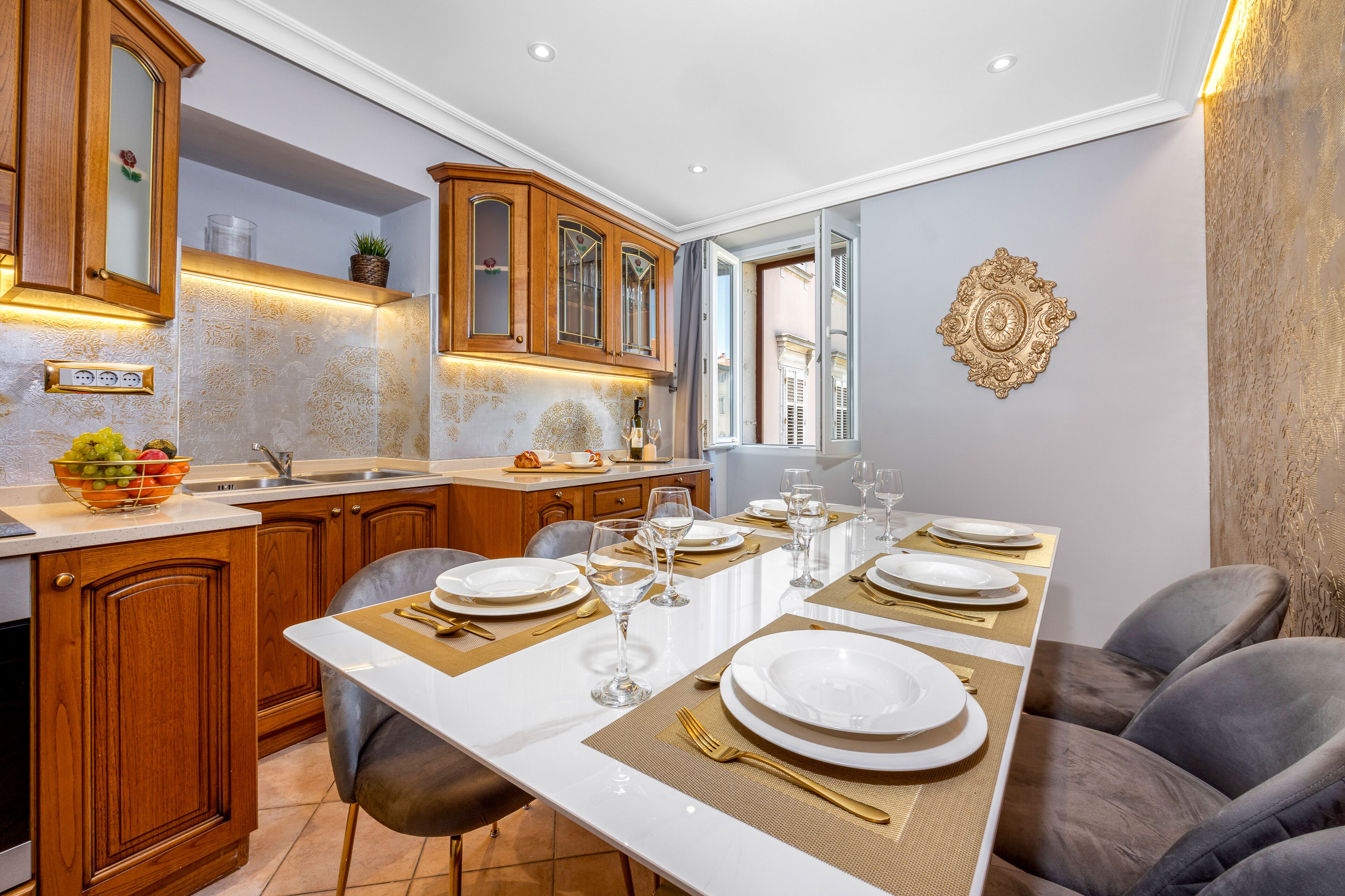 A modern kitchen with wooden cabinets, a white countertop, and a dining area set for six with a long table, chairs, and a chandelier.