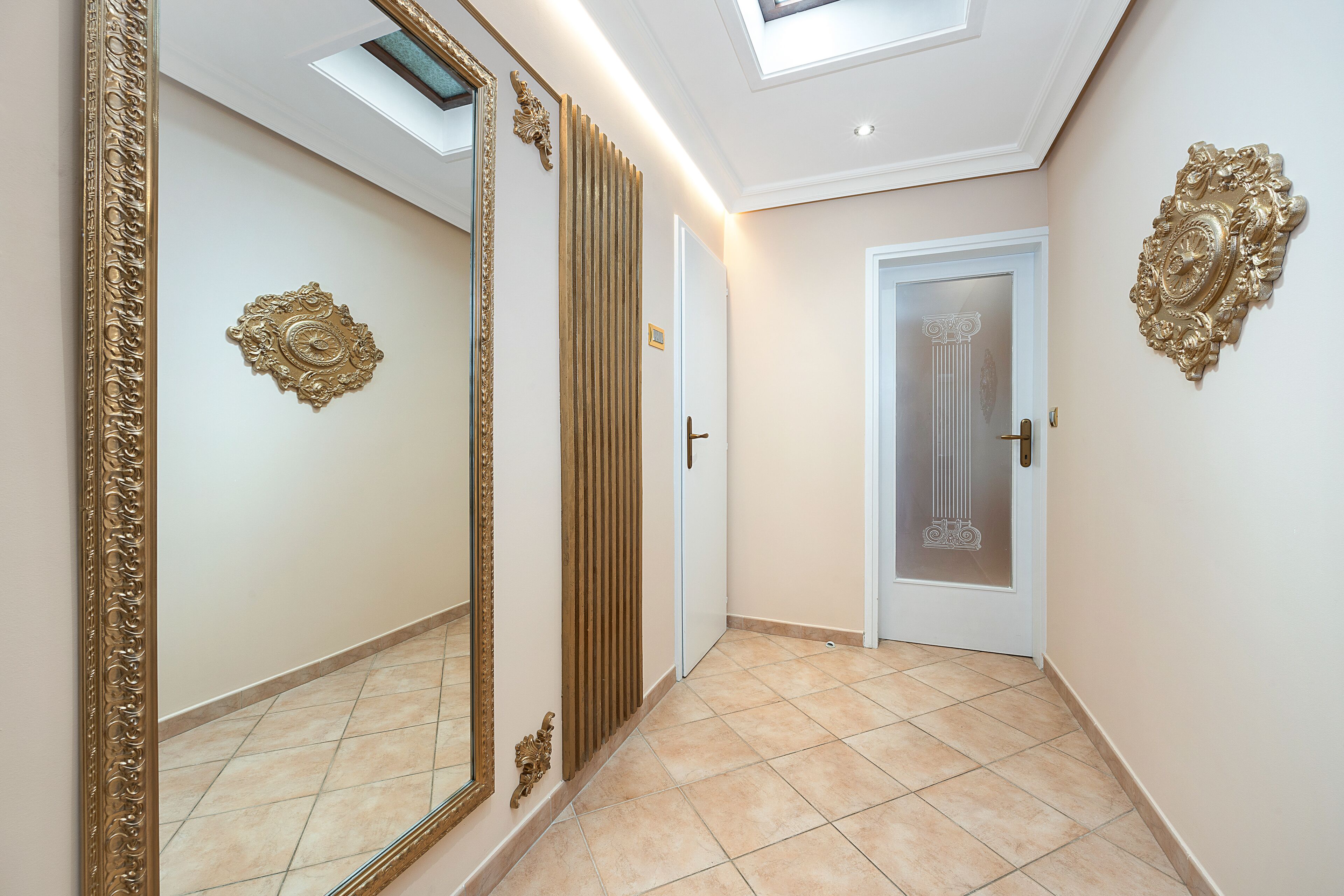 A hallway with a large ornate mirror, a door with a decorative glass panel, and a ceiling skylight.