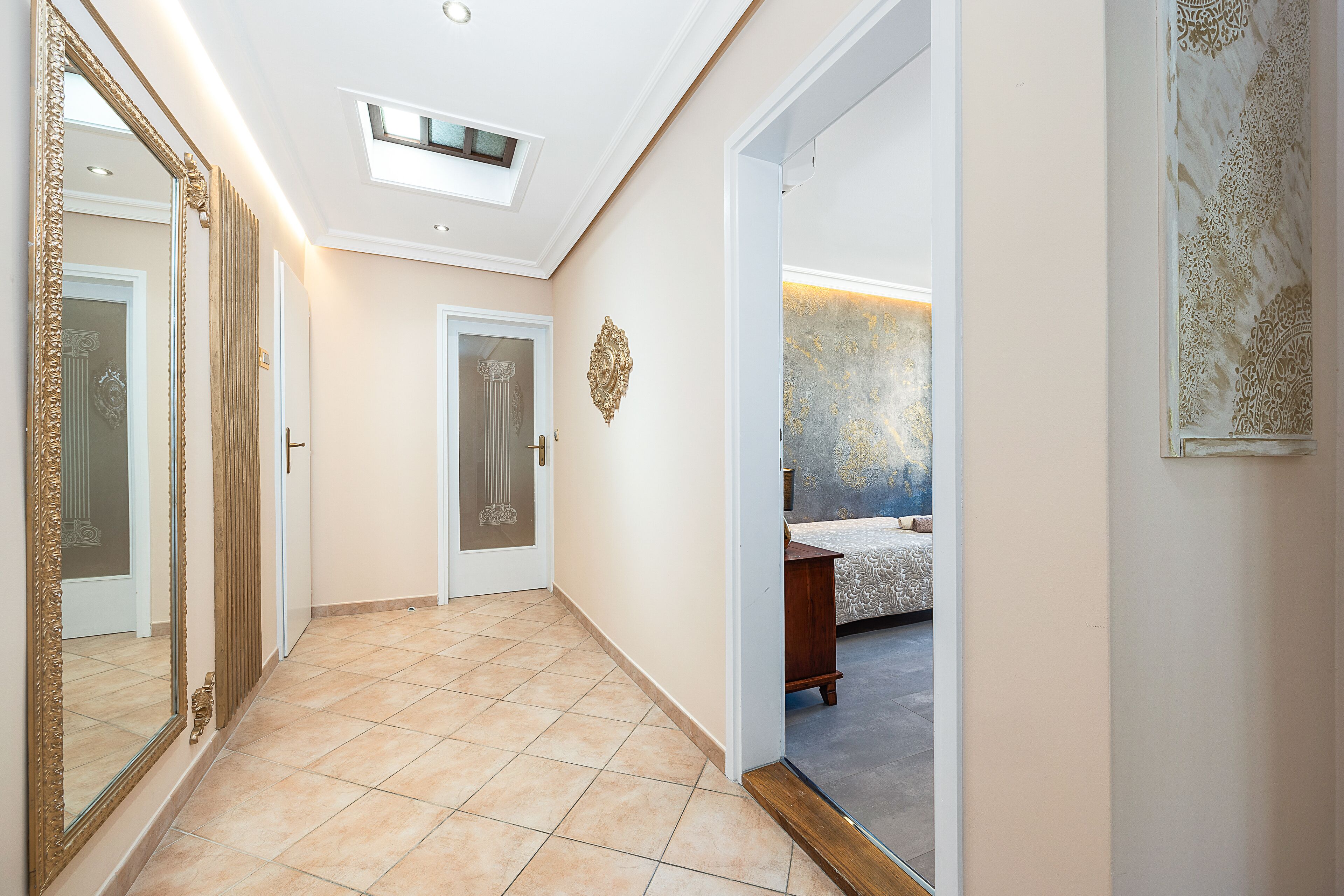 A hallway with a large mirror, a door with a decorative handle, and a view into a bedroom with a patterned bedspread.