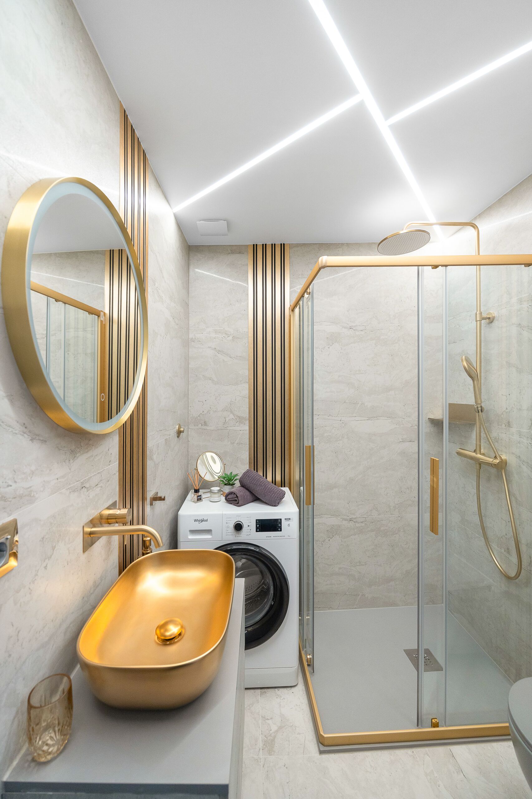 Une salle de bain moderne avec un lavabo doré, un miroir rond, une douche en verre et un lave-linge.