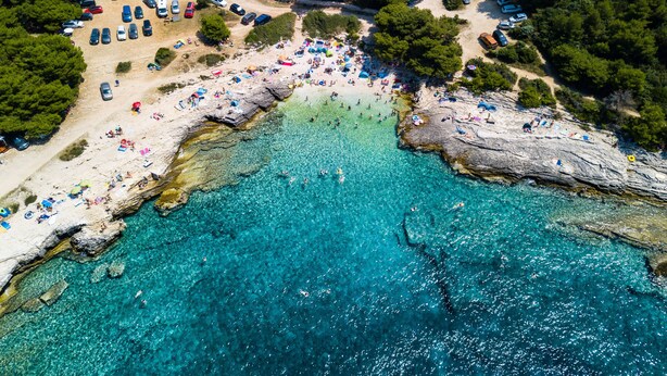 An aerial view of a beach with clear turquoise waters, rocky formations, and numerous beach umbrellas and sun loungers. - Augustus Forum View (Pula)
