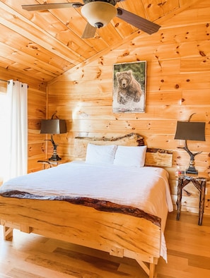 Ein Schlafzimmer in einer Holzhütte mit einem Bett, zwei Nachttischlampen, einem Deckenventilator und einem Bärenbild an der Wand.