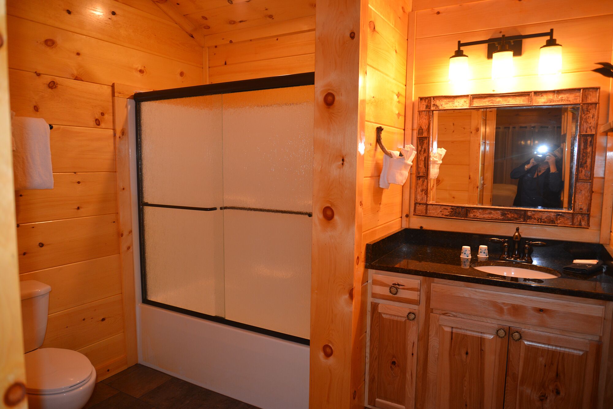 A wooden cabin bathroom with a toilet, shower, sink, and mirror.