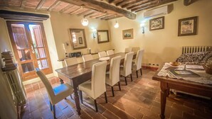 Un espace repas avec une longue table en bois et des chaises blanches, un sol en briques et un plafond rustique avec des poutres apparentes.