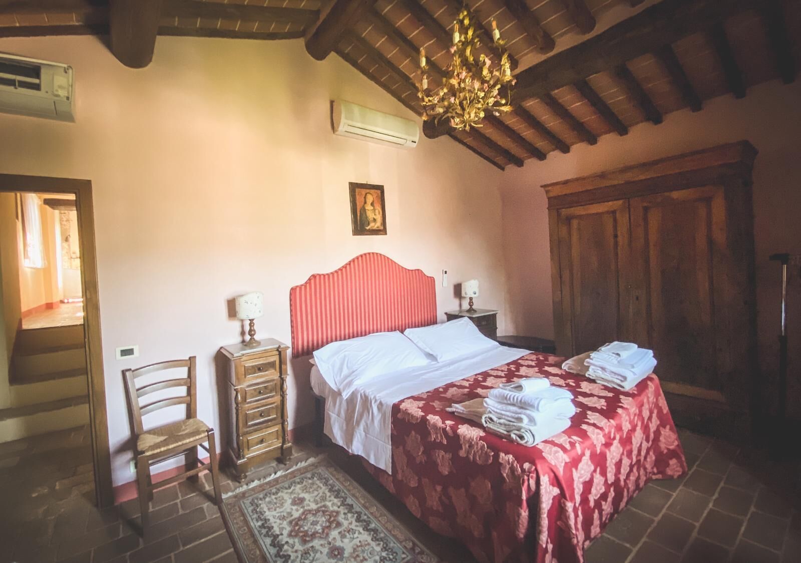 A bedroom with a red-striped headboard bed, a wooden nightstand with a lamp, a chair, a large wooden wardrobe, and a red floral bedspread.