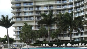 Una piscina con un diseño de baldosas hexagonales, una barandilla blanca y sillas de descanso. Al fondo, un edificio de varios pisos con balcones y palmeras.