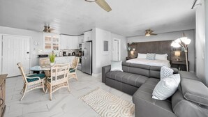Un salon moderne et bien éclairé, avec un canapé gris, un coin repas doté de chaises en osier, une cuisine aux meubles blancs et un ventilateur de plafond.