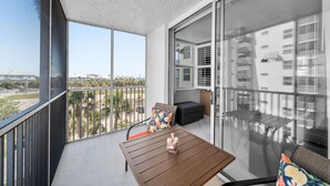Un balcon avec une table et des chaises en bois, un canapé et une porte vitrée donnant sur un autre balcon d’où l’on peut admirer les bâtiments et un ciel dégagé.