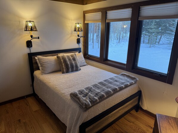 A bedroom with a bed, two wall-mounted lamps, a window with blinds, and a view of a snowy forest.