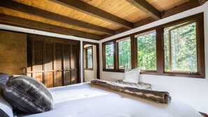 Un dormitorio con una cama grande, vigas de madera en el techo y vistas a un bosque por las ventanas.