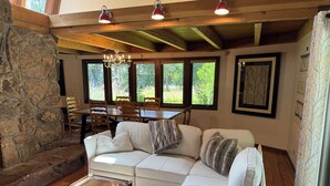 Una sala de estar acogedora con una pared de piedra, un sofá blanco, una mesa de centro de madera y una zona de comedor con sillas y mesa de madera.