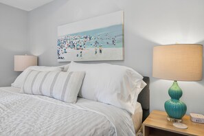 Quarto com cama, mesa de cabeceira, abajur e um quadro com tema de praia na parede.