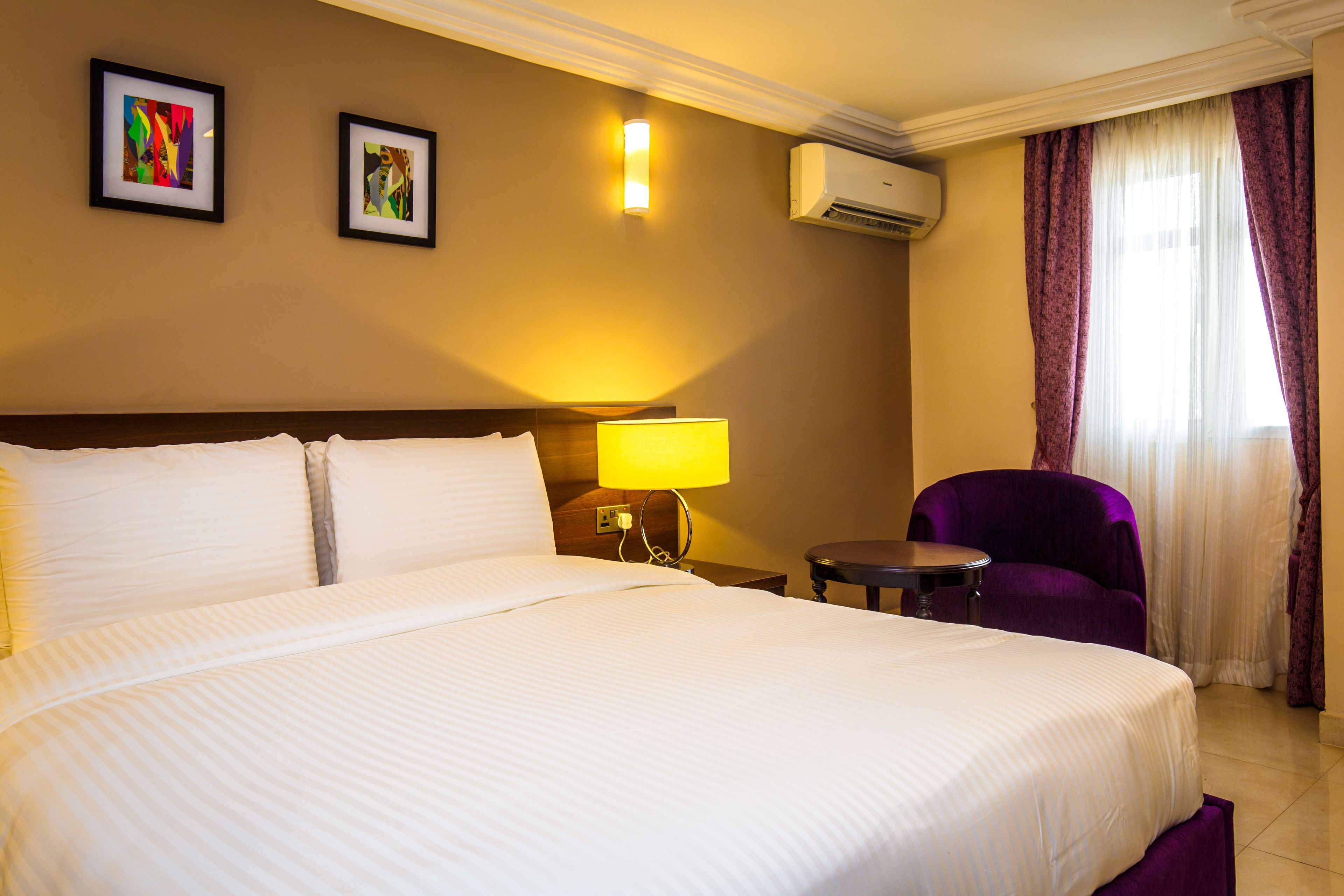 A hotel room with a large bed, a small table, a purple armchair, and two framed artworks on the wall.