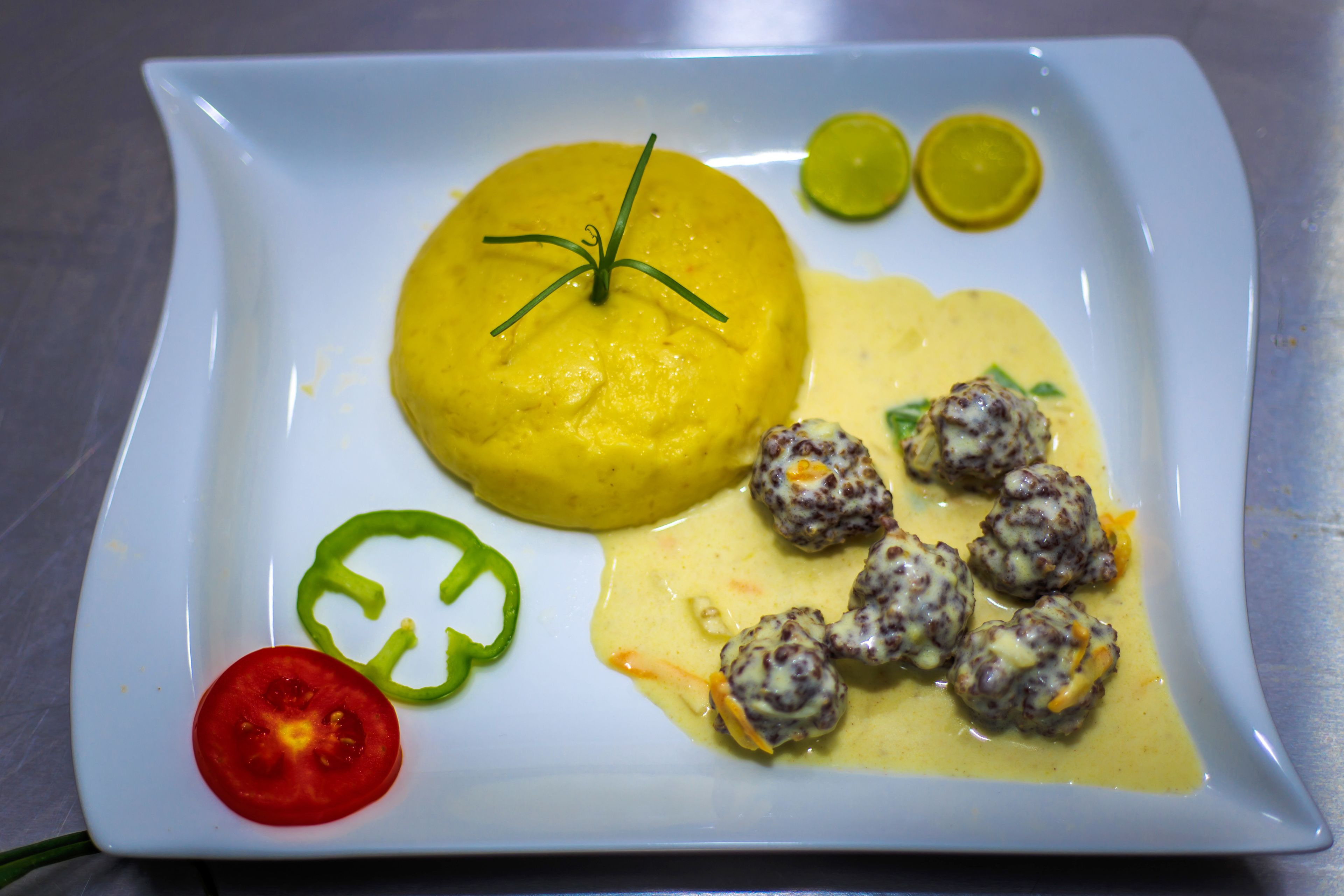 Ein quadratischer Teller mit gelbem Püree, Hackbällchen, einer Tomatenscheibe und einer Scheibe grüner Paprika.