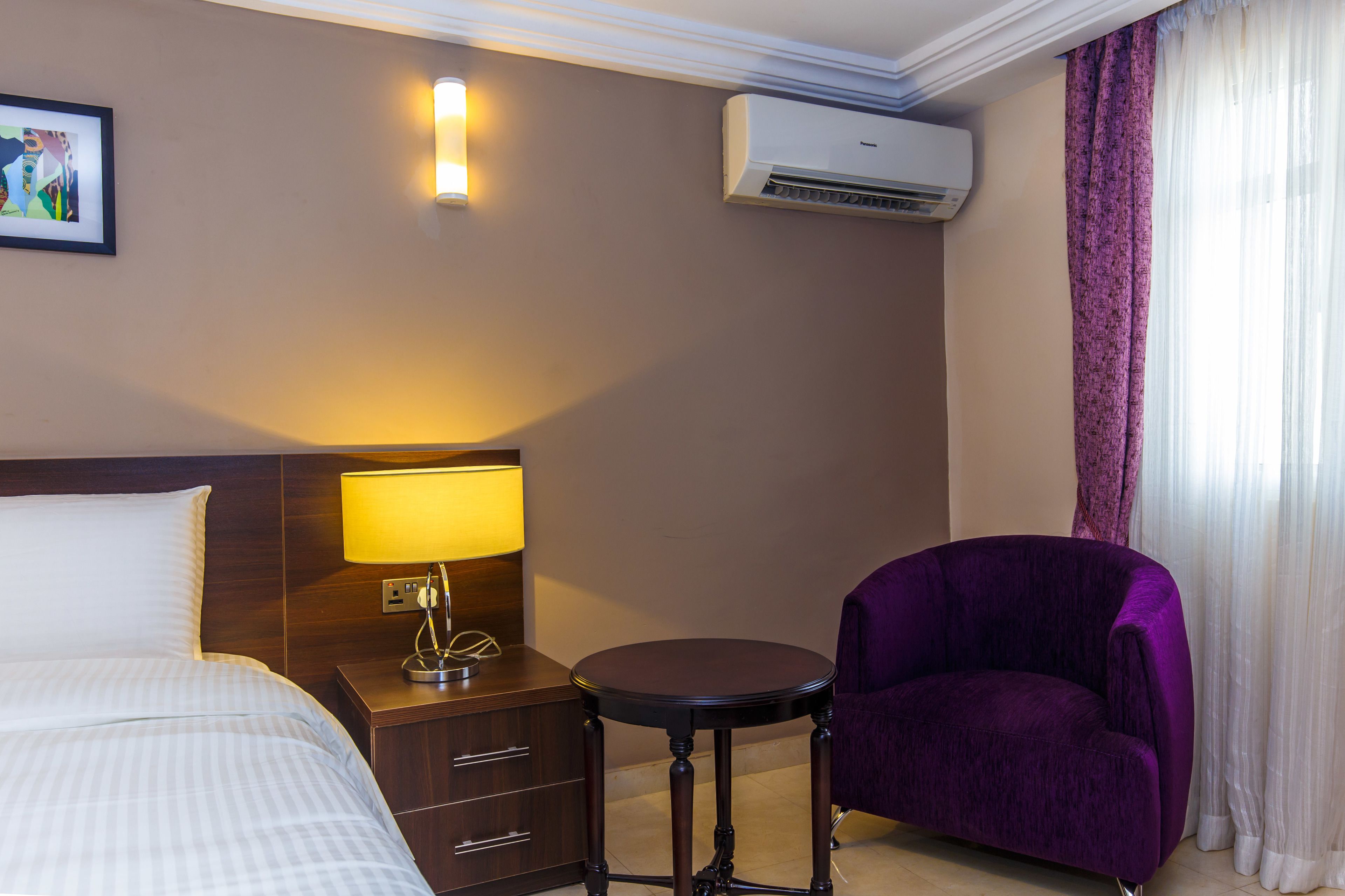 A hotel room with a bed, a nightstand with a lamp, a purple armchair, a small round table, an air conditioner, and a window with sheer curtains.