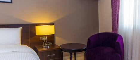A hotel room with a bed, a nightstand with a lamp, a purple armchair, a small round table, an air conditioner, and a window with sheer curtains.