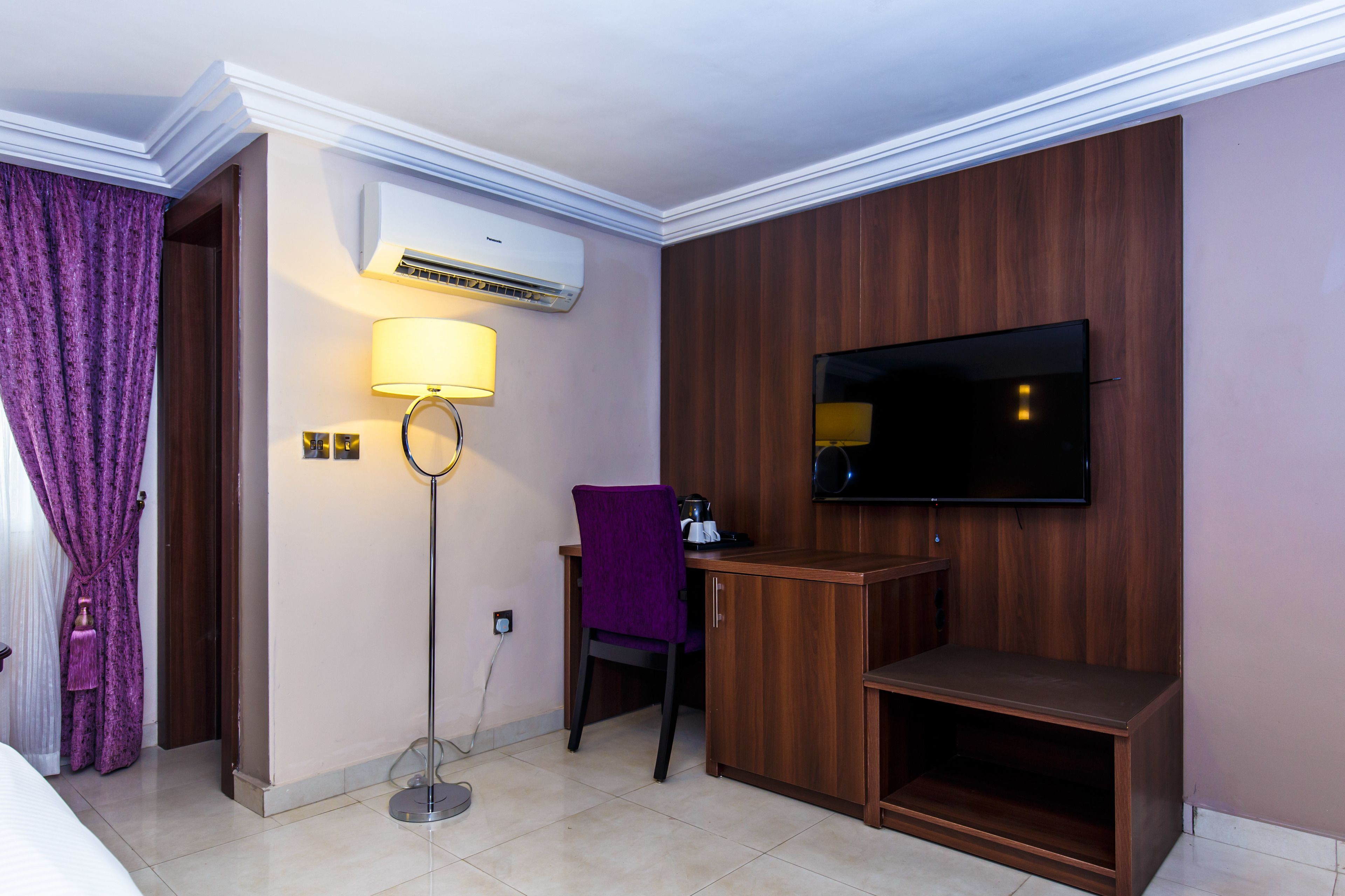 A hotel room with a bed, a desk with a chair, a TV mounted on the wall, an air conditioning unit, and a floor lamp.