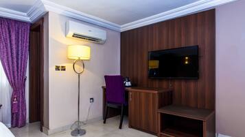 Habitación de hotel con cama, escritorio con silla, televisor montado en la pared, equipo de aire acondicionado y lámpara de pie.