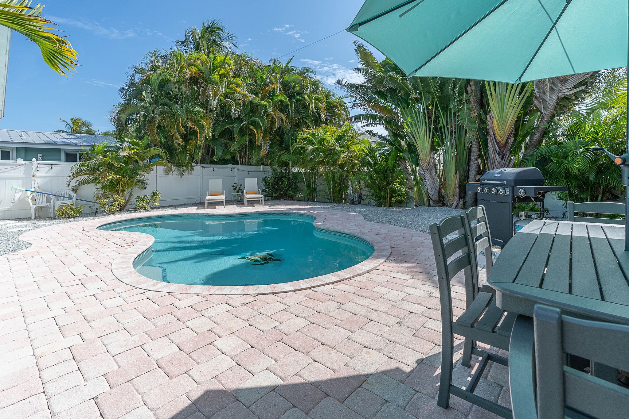 A swimming pool with a curved edge, surrounded by a brick patio. There are chairs and a table under a green umbrella.