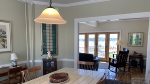 Ein Essbereich mit einem großen Tisch, Stühlen, einer hängenden Leuchte, einem Fenster mit dekorativen Fensterläden und Blick durch Glastüren auf eine Terrassenfläche.