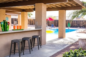 An outdoor bar area with a pool, bar stools, and a wooden fence in the background. - Tropical Paradise! Walking distance to Bridge/Channel! Huge Yard & Heated Pool (Lake Havasu City)