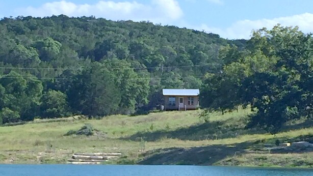A house with a blue roof, surrounded by a forested hillside and a body of water. - Papa's Palace at Stacy Ranch, cabin on 120 acres between Ingram & Hunt, TX (Ingram)