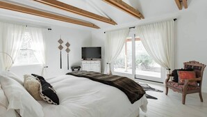 Ein Schlafzimmer mit einem großen Bett, einem Fernseher, einem Holzsessel und Blick ins Freie.