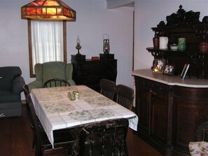 Un coin repas avec une table recouverte d’une nappe à motifs floraux, entourée de chaises en bois. Une lampe en vitrail est suspendue au-dessus de la table, et un buffet en bois orné d’objets décoratifs se trouve à droite.