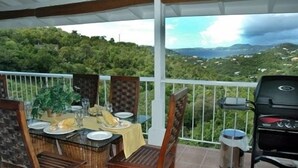A covered outdoor dining area with a table set for two, a grill, and a view of a hilly landscape.