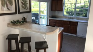 A kitchen with a large island, bar stools, wooden cabinets, a window with a view of greenery, and framed artwork on the wall.
