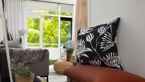 A modern living room with a sofa, a red bench, a large window with a view of greenery, and a decorative pillow. - St Martini 416 (Cape Town)