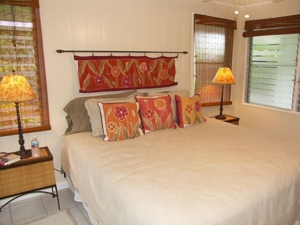 A bedroom with a large bed, floral-patterned pillows, two bedside lamps, a wooden nightstand, and a window with blinds. - MONTHLY DISCOUNT! 3Bed/2Ba House - 250 Steps to the Beach! (Kailua)