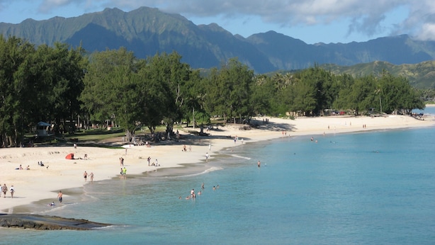 Ein Strand mit klarem, blauem Wasser, Sandküste und Bergen im Hintergrund.